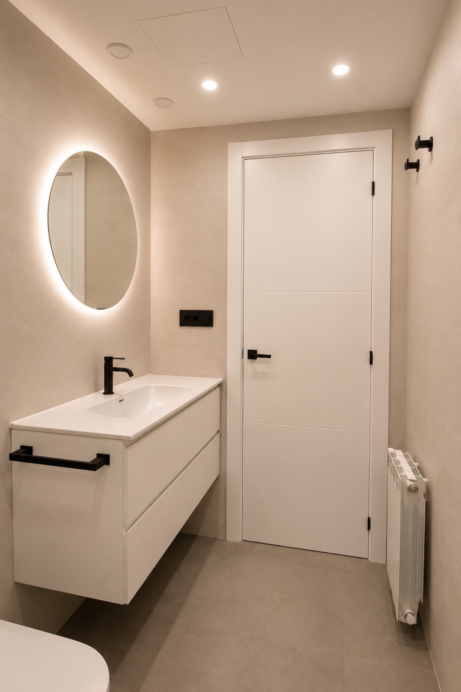 Marfil y negro. Un mueble claro, espejo circular y accesorios negros en un baño compacto. La ducha queda separada por una mampara transparente.