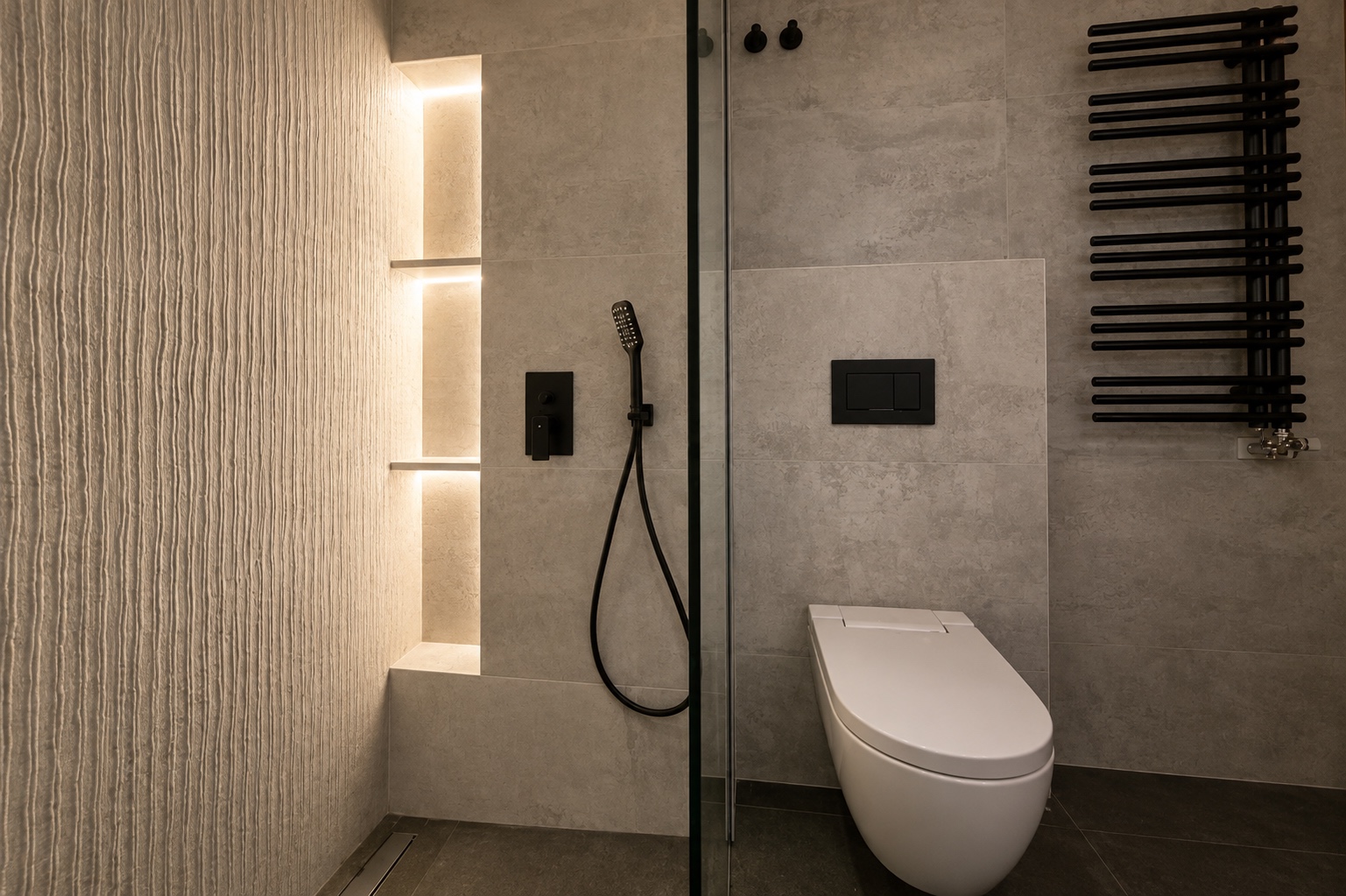 Relieve y luz. Una pared texturizada y una hornacina iluminada delimitan la ducha. Los accesorios negros contrastan con el revestimiento gris.